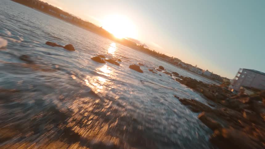 A fast-paced FPV drone flight skims just above the ocean waves crashing against a rocky shoreline during a brilliant golden sunset