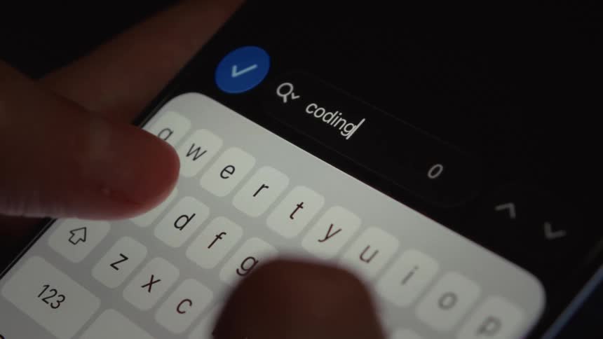 A close-up of fingers typing 'coding' into a search bar on a smartphone screen in a dimly lit room