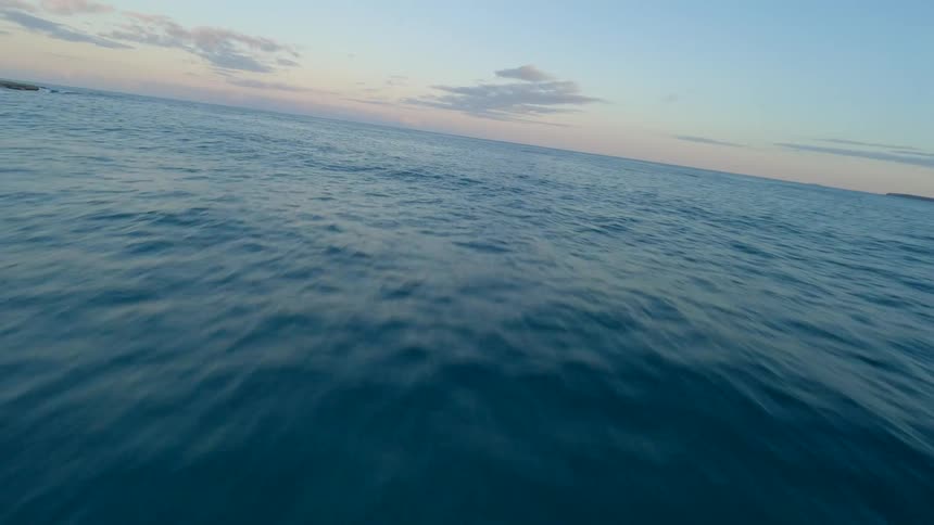 A fast FPV drone shot flying low and close over the deep blue ocean water at sunset