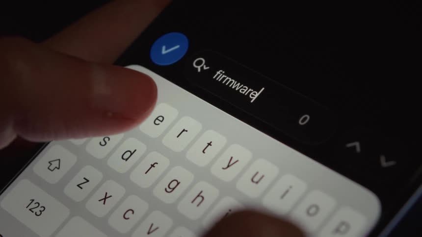 A close-up view of fingers typing the word 'firmware' into the search bar of a smartphone in a dark room