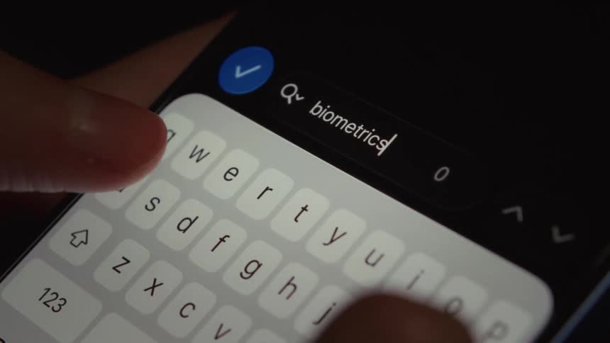 A person types the word 'biometrics' into the search bar on a smartphone in a dark environment