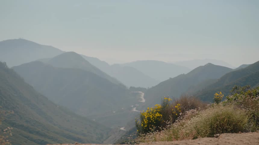 A hazy, scenic view of a vast mountain range reveals a winding road through the deep valley below