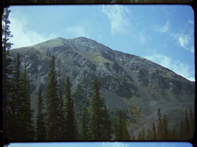 A vintage film view of a rugged mountain peak rising above a pine forest against a bright blue sky