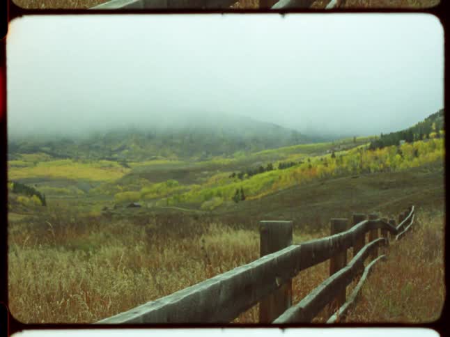A vintage film effect with a red light leak fades to reveal a rustic wooden fence overlooking a foggy autumn valley