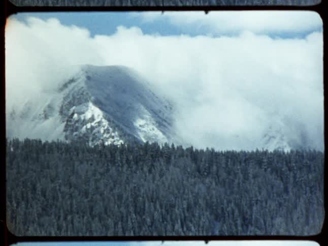 Vintage 8mm film captures a zoom-in shot of a majestic snow-covered mountain peak surrounded by clouds over a vast evergreen forest