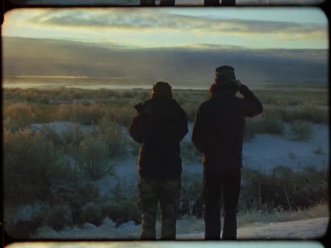 A vintage 8mm style shot captures two friends filming the golden sunrise over a vast, snowy winter landscape