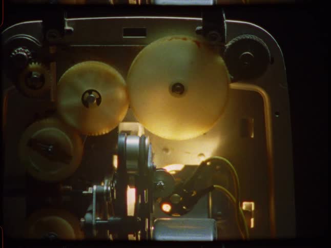 A slow zoom-in shot captures the internal mechanisms and spinning gears of an old 8mm film projector, illuminated by its warm lamp
