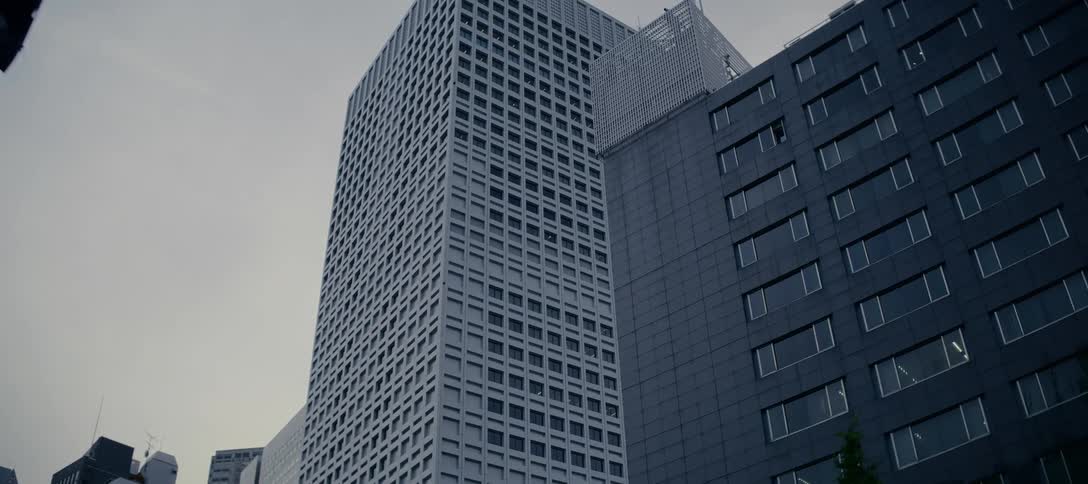 A red traffic light turns green against a backdrop of imposing, monochromatic skyscrapers in a bleak urban cityscape