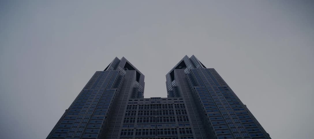 A slow tilt-down reveals the imposing facade of the twin towers of the Tokyo Metropolitan Government Building against a gray sky