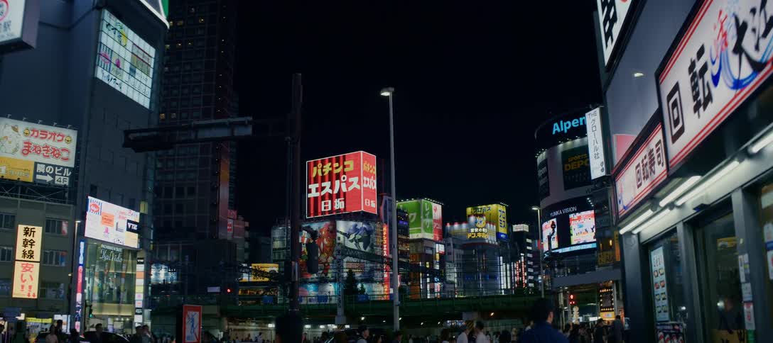 Vibrant neon signs and billboards illuminate a bustling street intersection in Shinjuku, Tokyo at night as pedestrians and trains pass by