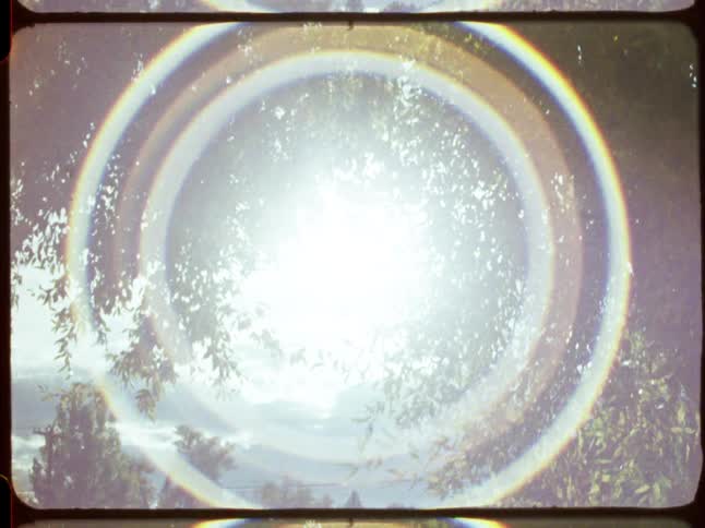 Sunlight streams through lush green tree leaves creating a dreamy circular rainbow lens flare in this authentic vintage-style footage