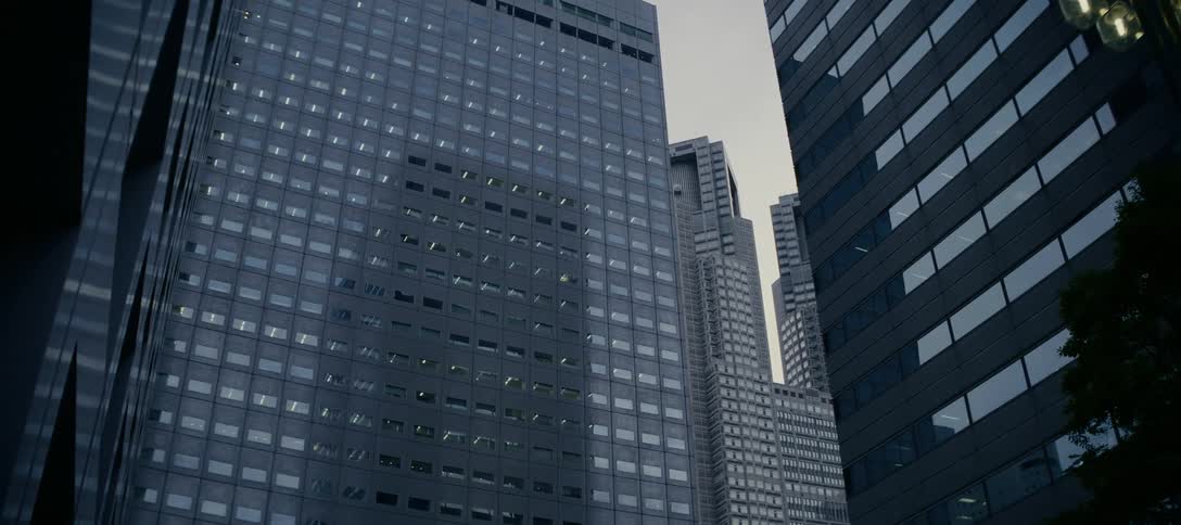 A slow push-in shot looks up at imposing modern skyscrapers in a city financial district under a gloomy, overcast sky