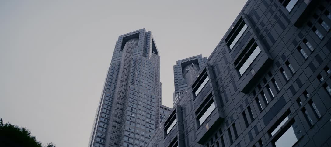 A slow, low-angle reveal of the iconic Tokyo Metropolitan Government Building from behind a tree against an overcast sky
