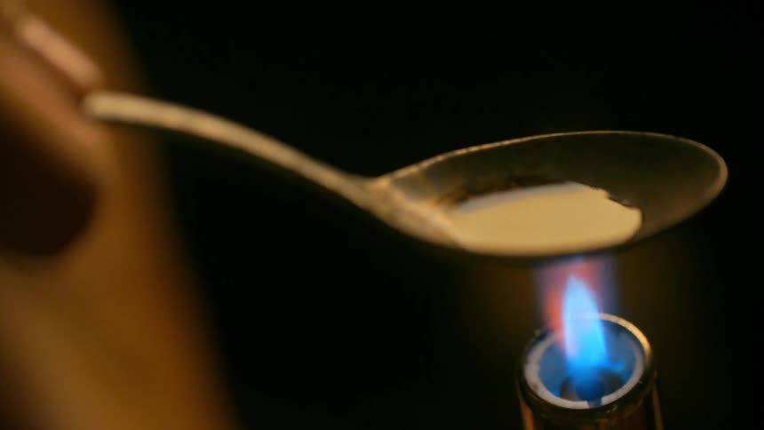 Extreme close-up of a person heating a powdered substance in a spoon with a lighter until it boils and turns into a dark liquid