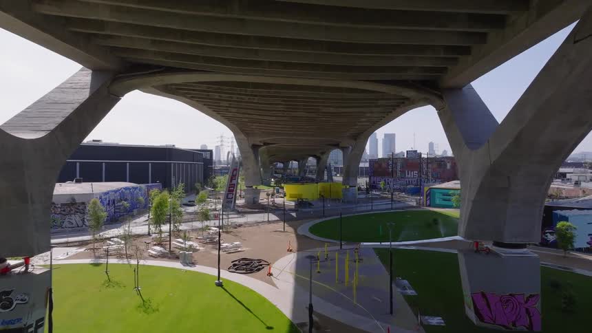 Slow aerial push-in shot under a massive concrete bridge revealing the construction of a new urban park in Los Angeles