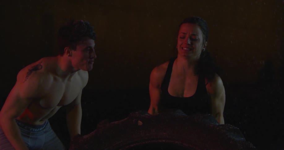 Man coaches a woman as she pushes a large tire over