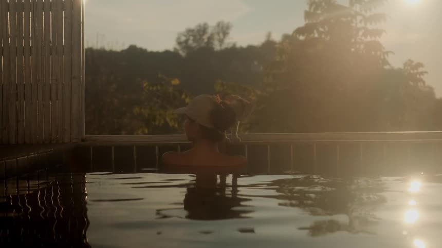 A woman relaxes in a pool at golden hour as her curious cat drinks from the edge and then playfully walks over her head