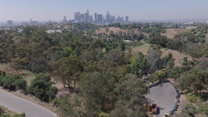 Drone footage flies over a wooded park with the iconic Downtown Los Angeles cityscape on the horizon