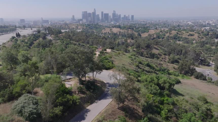 A slow aerial push over the winding roads and green hills of a large park with the Los Angeles city skyline in the background