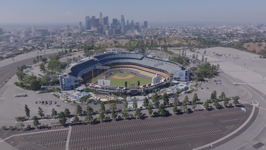 A slow aerial dolly shot pushes in on Dodger Stadium, surrounded by an empty parking lot with the Los Angeles city skyline in the background