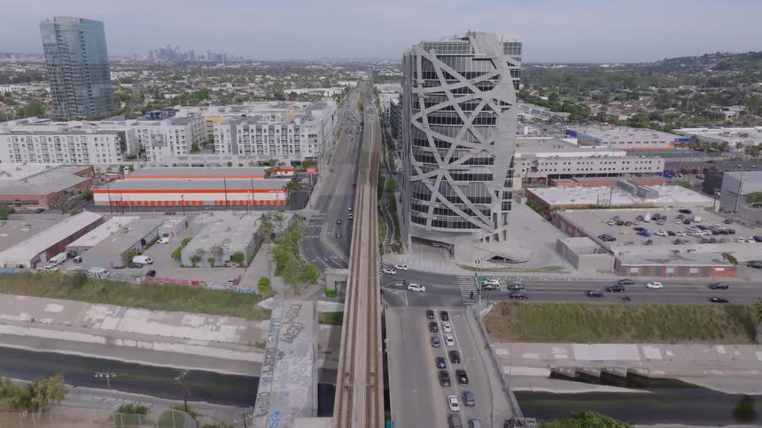 An aerial view flies over the Metro E Line train tracks toward the distinctive 'Wrapper' building in Culver City, California