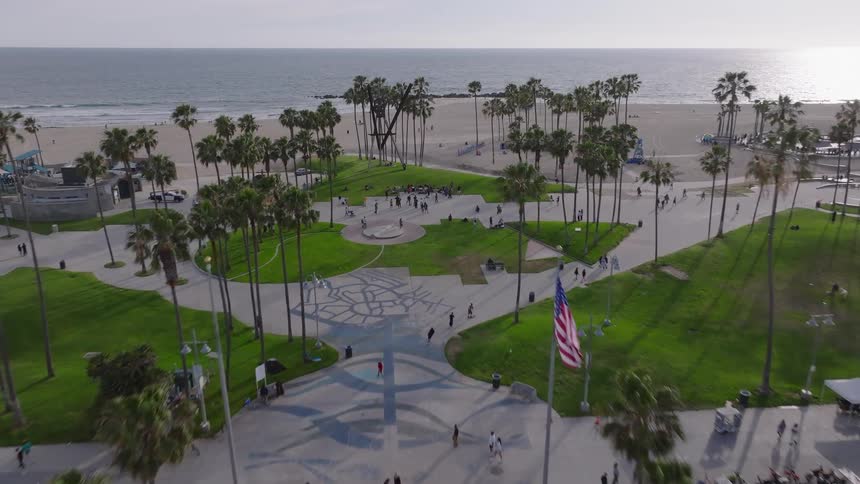 An aerial drone shot flies down a street in Venice Beach, California, revealing the boardwalk, palm trees, and the Pacific Ocean at sunset