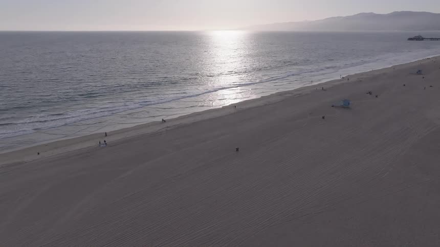 A tranquil aerial view captures the sun reflecting on the ocean as it moves along a vast, sandy beach with scattered people and lifeguard towers