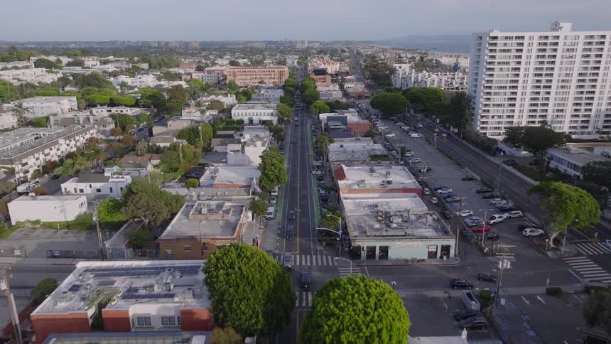 An aerial view flies down a picturesque, tree-lined street in a bustling coastal city towards the ocean