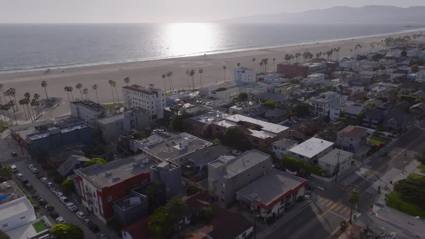 An aerial view pans slowly across the Venice Beach cityscape in California, showing the vibrant neighborhood and the Pacific Ocean coastline at golden hour