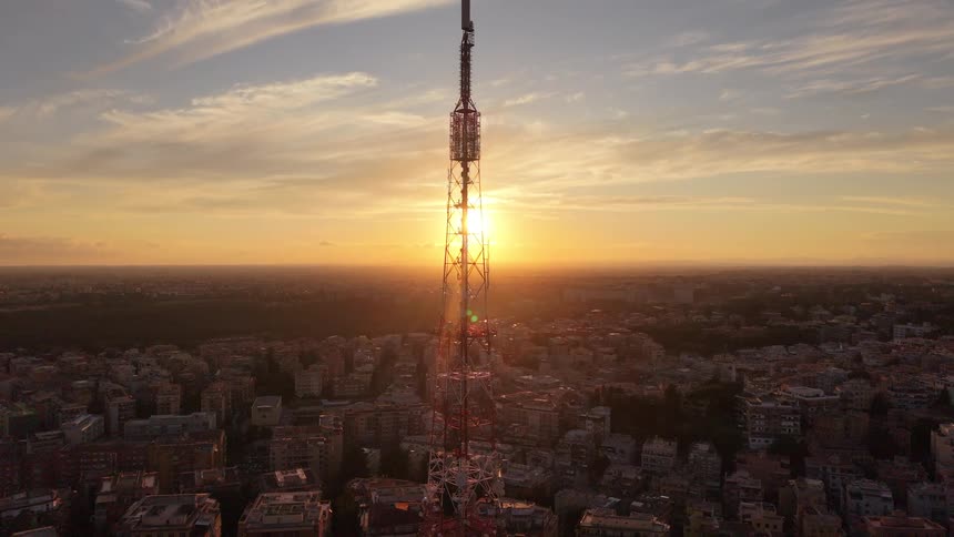 Aerial view pulling back from a large communications tower silhouetted against a beautiful city sunset