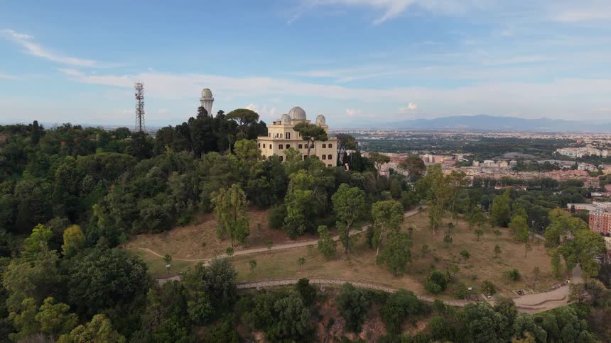 An aerial view reveals the majestic Astronomical Observatory of Rome perched atop the lush Monte Mario hill, with the sprawling city skyline in the background