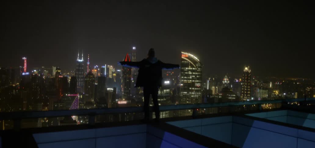 A man stand on a rooftop overlooking a city at night and lifts his arms