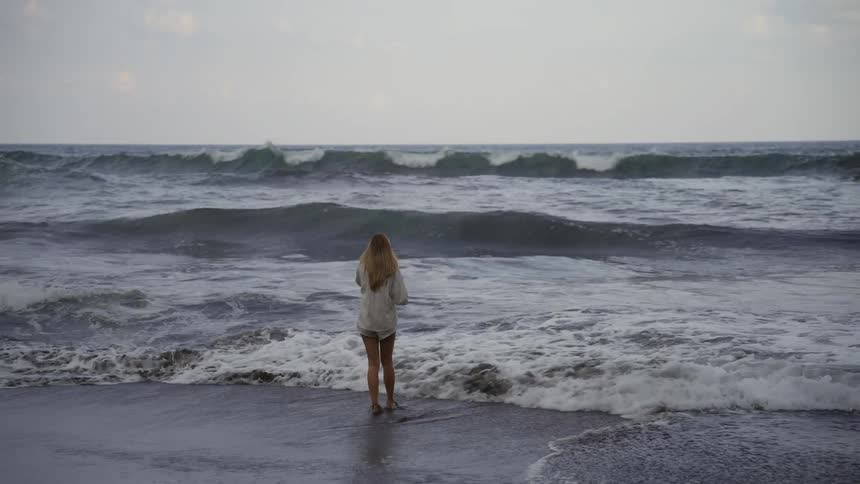 A woman stands in the shallow surf of a stormy ocean, watching the waves crash in slow motion on a cloudy day