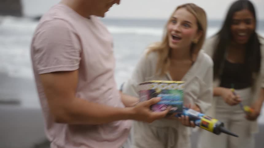 A young man brings a large box of fireworks to the beach, happily sharing them with his excited friends