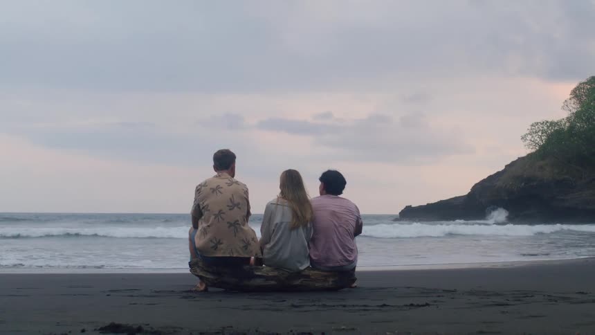 Three friends sit together on a driftwood log on a dark sand beach, talking and watching the ocean on a cloudy evening