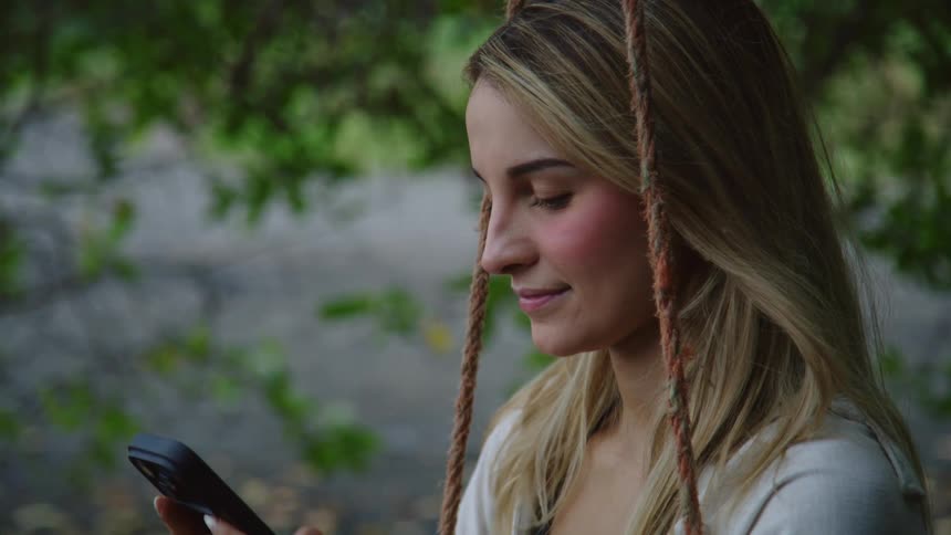 A young woman with a serene smile scrolls on her smartphone while relaxing on a rope swing outdoors