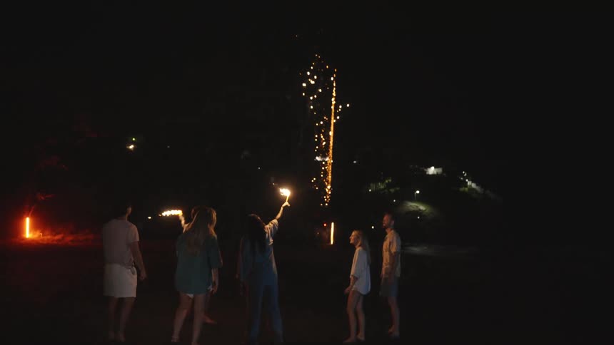 A group of friends stands together on a dark beach at night, holding sparklers and watching fireworks explode in the sky