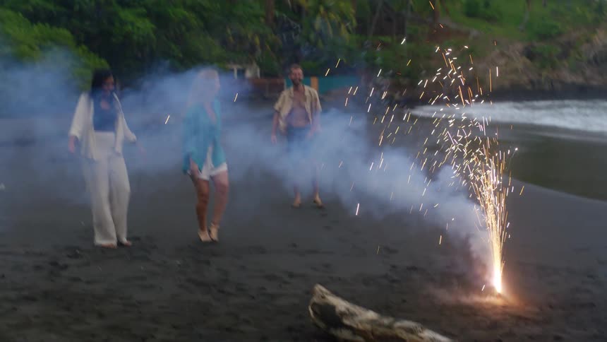 Friends celebrate with a firework on a tropical black sand beach as one man performs a fun somersault
