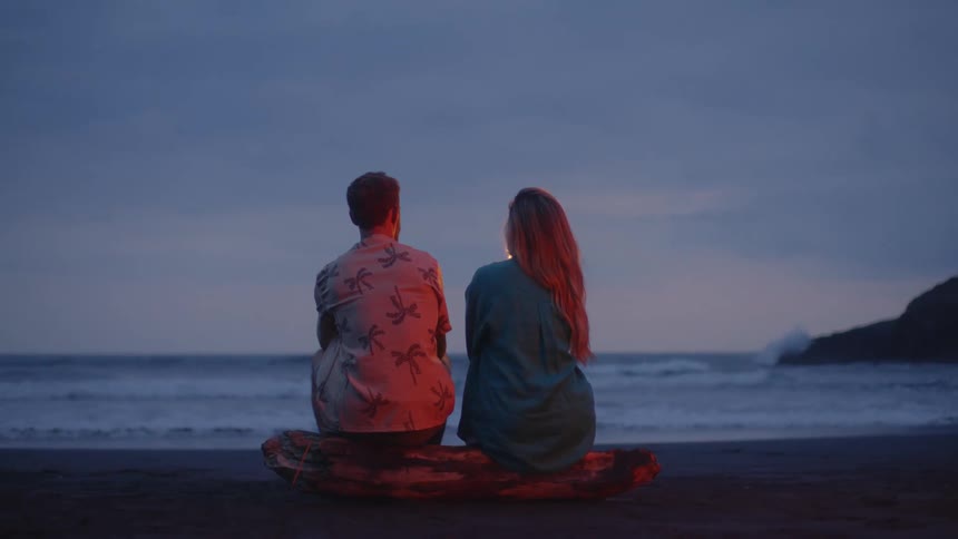 A young couple sits on a log on the beach at twilight, enjoying a romantic moment with a sparkler while watching the ocean