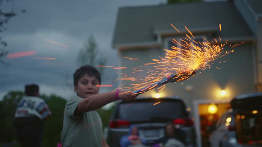 A young boy smiles while holding a large firework that showers brilliant golden sparks during a suburban Fourth of July celebration in slow motion