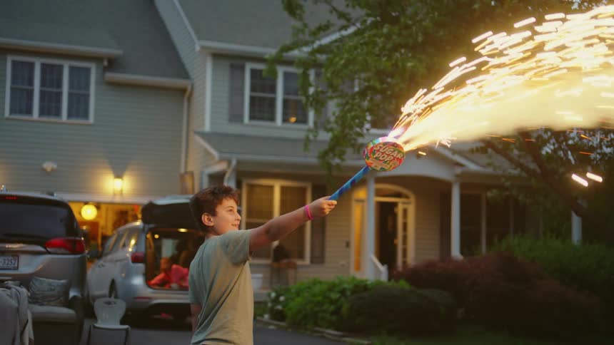 A young boy's face glows with excitement as he holds a large sparkling firework during a suburban Fourth of July celebration at dusk