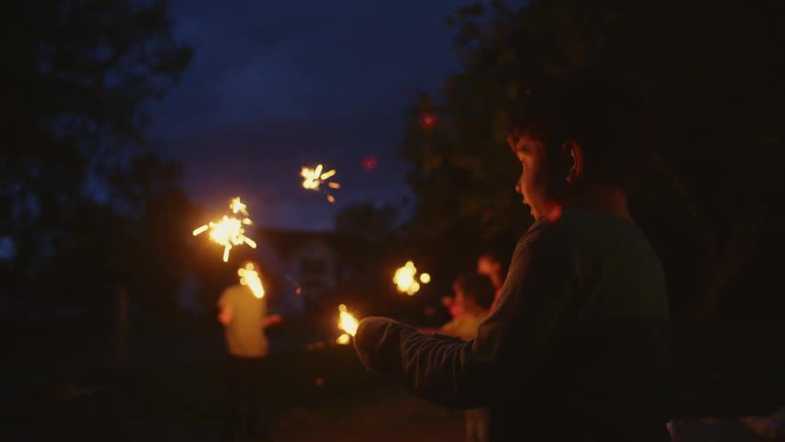 In slow motion, a young boy's face is illuminated with joy by a sparkler as he celebrates with friends at night