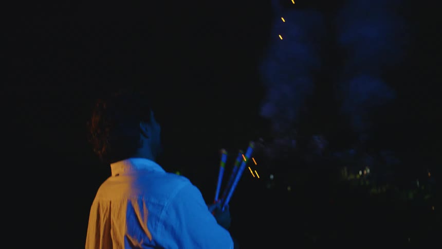 A man seen from behind launches colorful Roman candle fireworks into the night sky, illuminating himself and the surrounding area