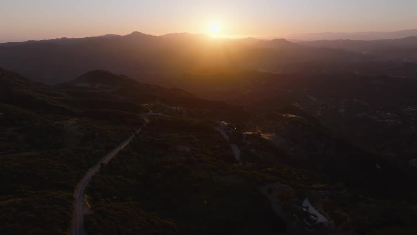 An epic aerial shot shows a winding road through a vast mountain landscape during a beautiful golden sunset
