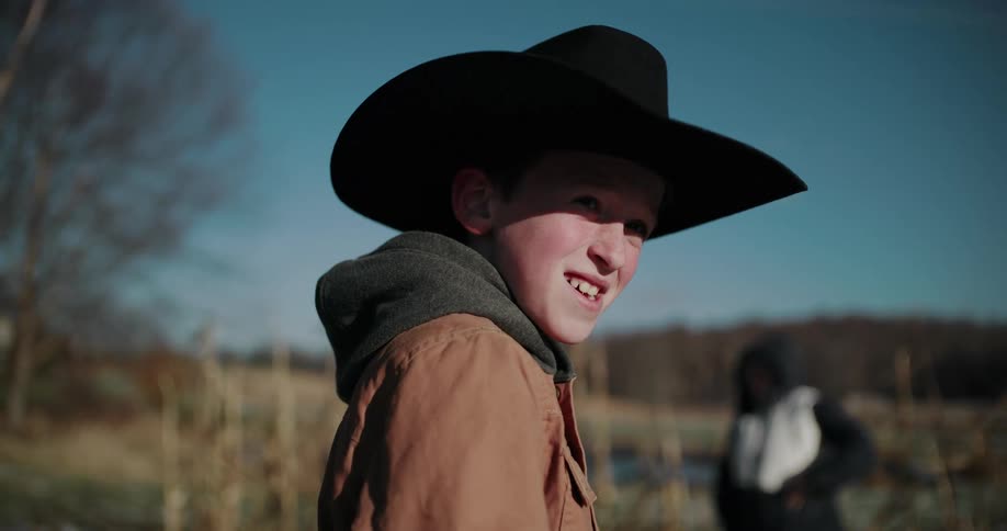 A smiling boy in a black cowboy hat with a feather turns to the camera on a sunny day in a golden field