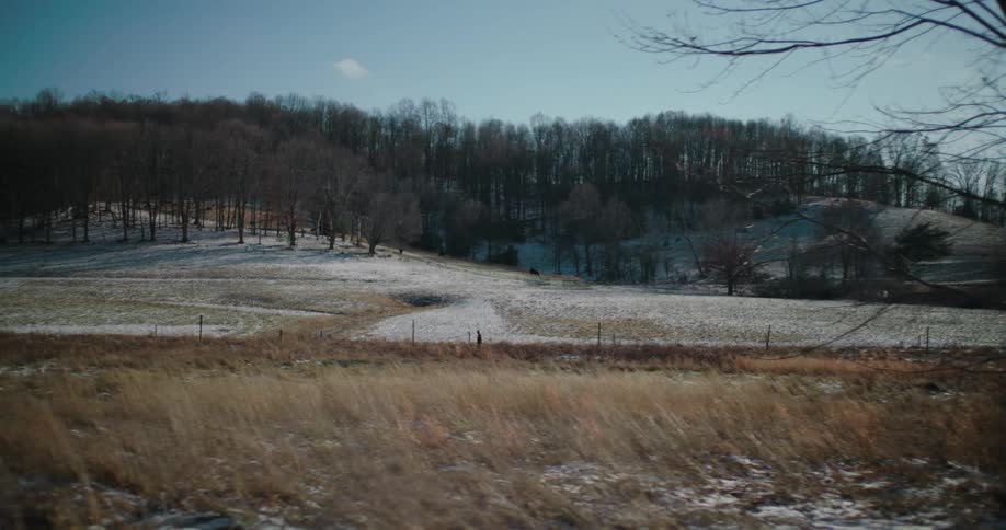 A tracking shot reveals a serene, snow-dusted rural landscape with rolling hills and bare trees on a sunny winter day
