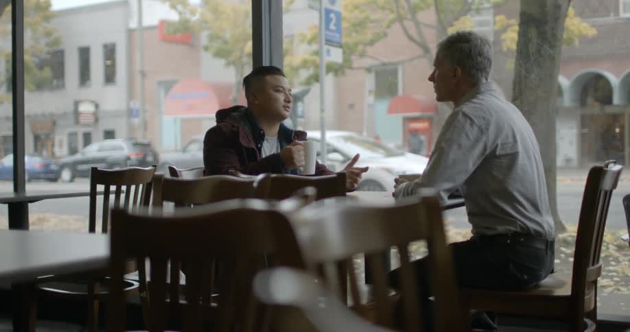 Two men sit at a table talking in a coffee shop
