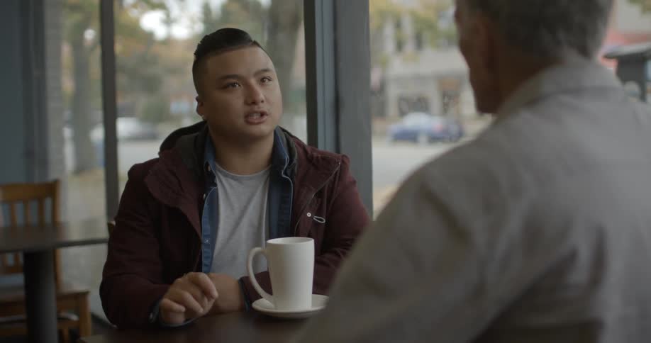 Two men sit at a table talking in a shop in front of a large window