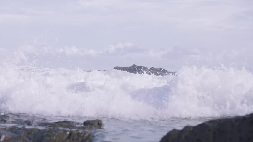 A powerful ocean wave crashes against a rocky shoreline and washes over the rocks in dramatic super slow motion