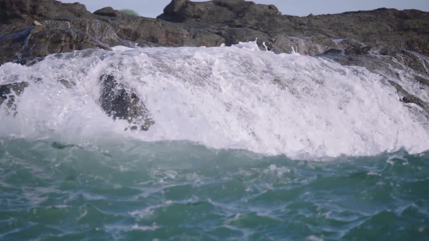 Extreme slow motion close-up of powerful ocean waves crashing over dark, jagged rocks on the coastline